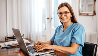 Female nurse in blue scrubs sitting at a desk, smiling, and typing on laptop doing clinical charting in a nursing home office